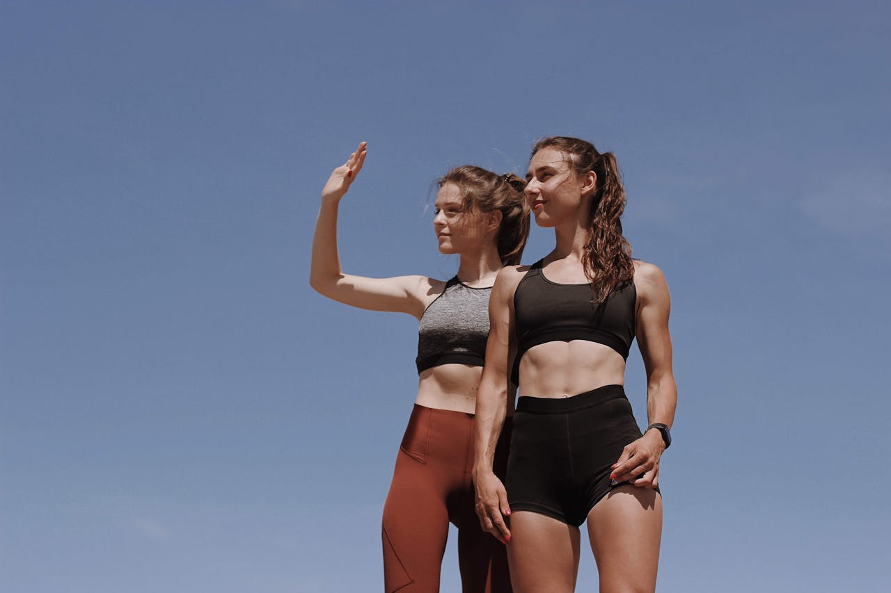 Two fit young women in workout attire posing confidently against a clear blue sky.